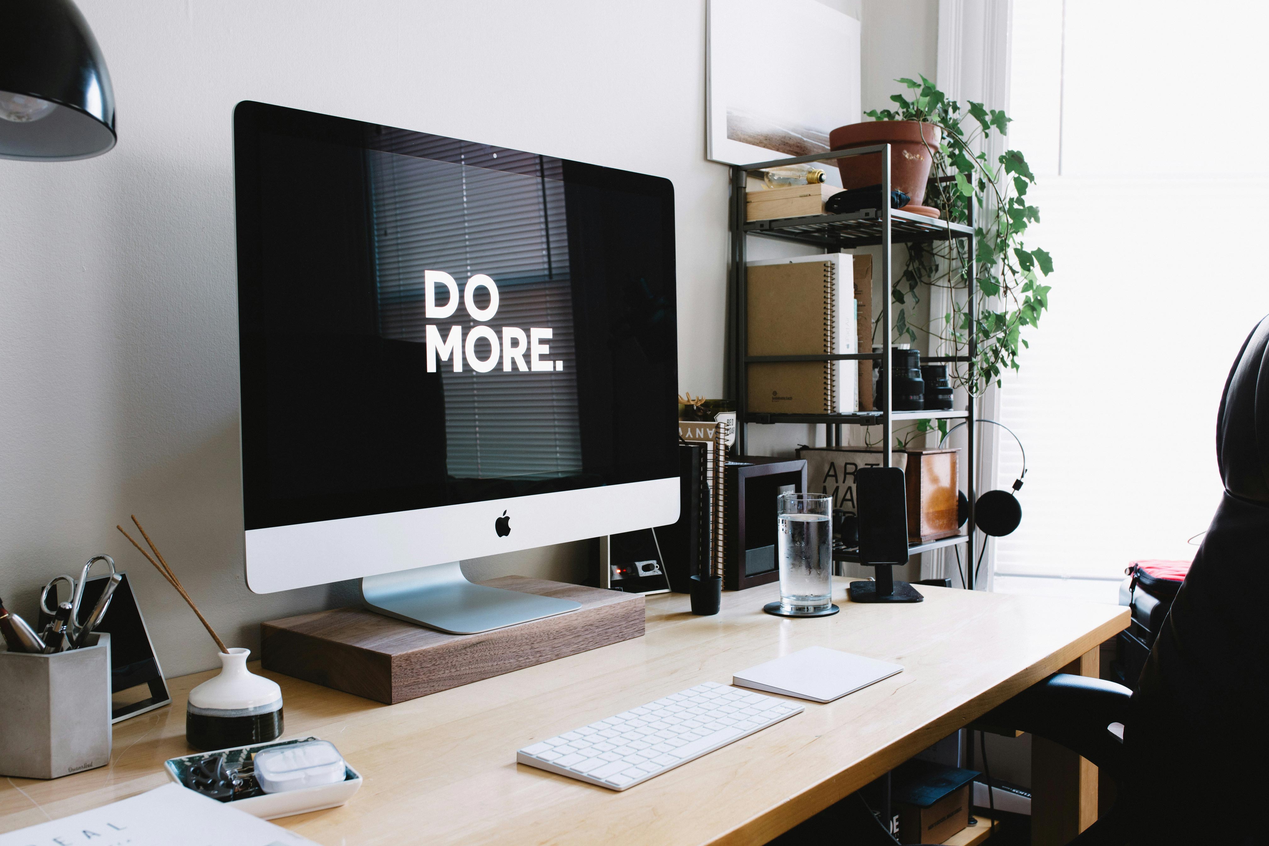 Moderner, aufgeräumter Arbeitsplatz mit iMac auf einem Holztisch. Auf dem Bildschirm steht in weißer Schrift „DO MORE.“ – umgeben von Pflanzen, Regalen, Notizbüchern und minimalistischer Büroeinrichtung.